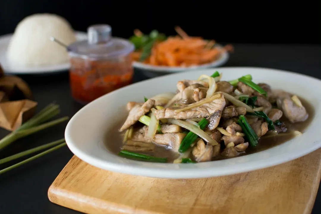 Sliced meat stir-fry with green onions on a white plate, with rice and chili sauce behind it