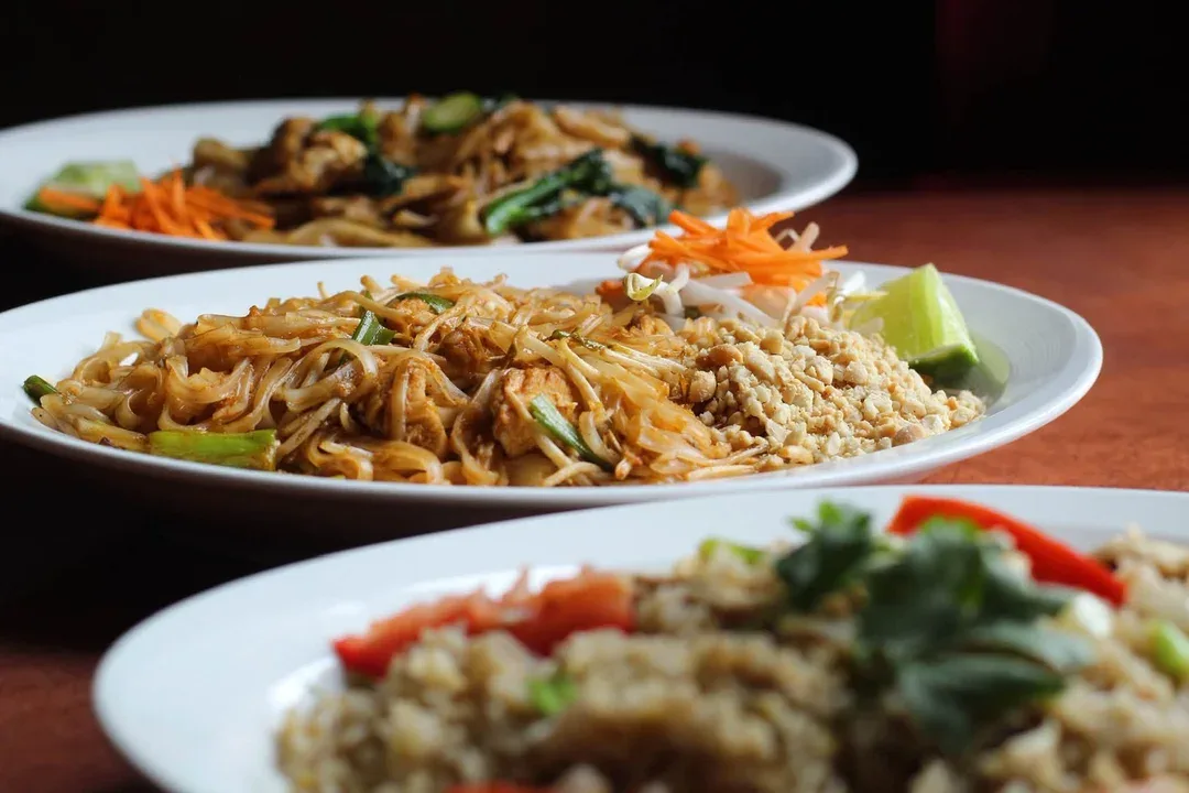 Three plates of Thai noodles and fried rice, with pad thai topped with peanuts and lime in front